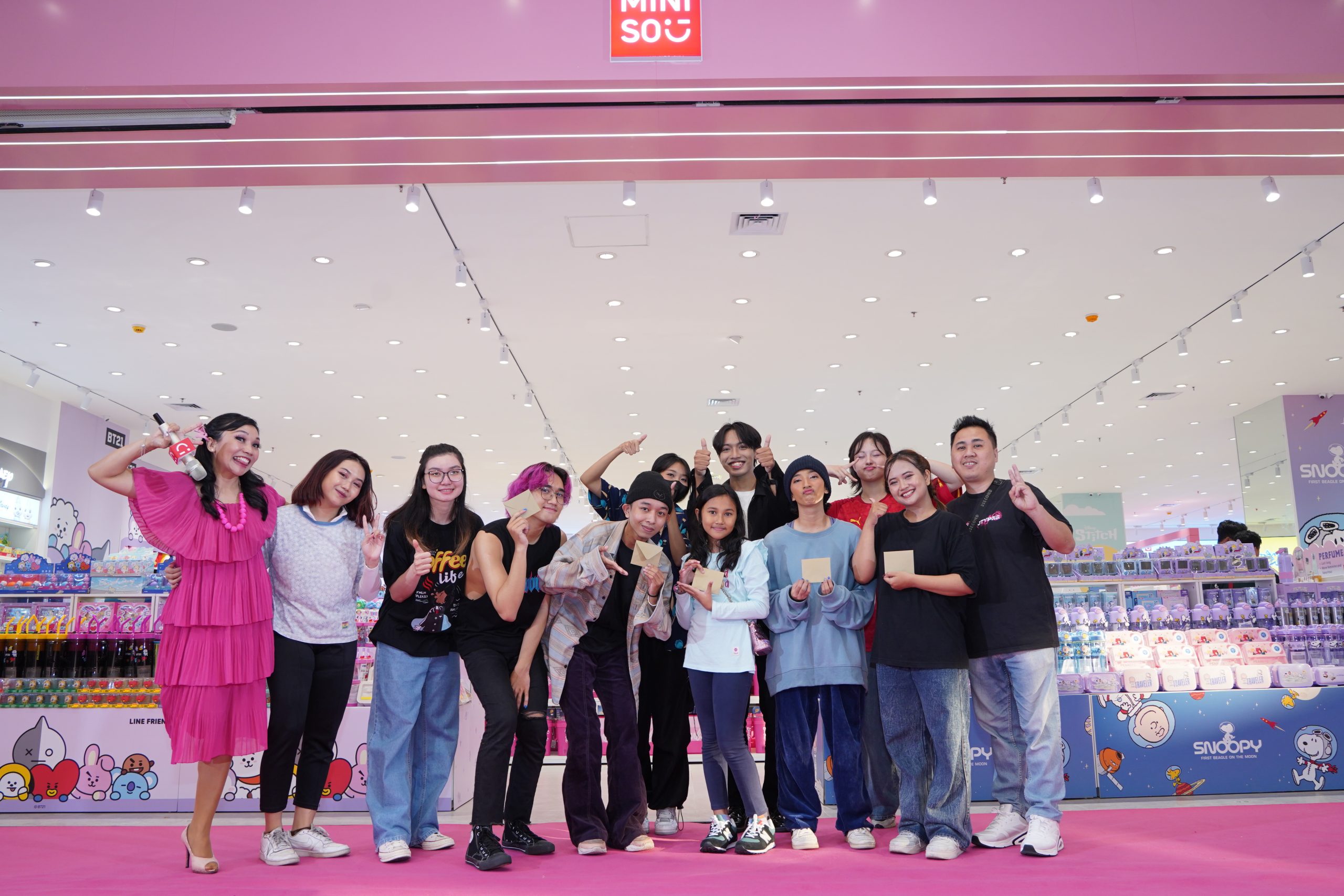 This image shows a group of people posing in front of a Miniso store. The store's entrance is decorated with bright, playful colors, primarily pink, and features merchandise that seems to be themed around popular characters, as suggested by the visible products on the shelves. The group includes a mix of people, likely staff or event participants, dressed in casual attire. They are smiling and making various hand gestures, showing their excitement and camaraderie. The atmosphere is lively and celebratory, typical of a grand opening event.