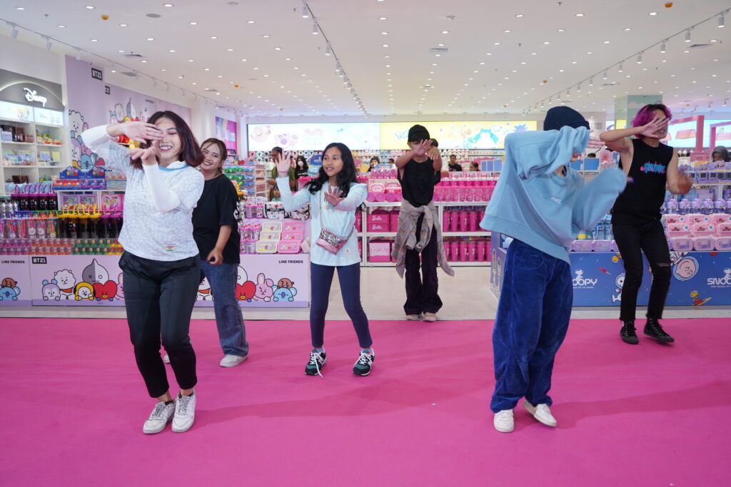 A group of young people perform a dance on a bright pink carpet inside the newly opened MINISO PINK store at The Park Semarang. The store is filled with colorful merchandise, including LINE FRIENDS, Barbie, and Snoopy items, with pastel decor and vibrant displays creating a lively atmosphere.