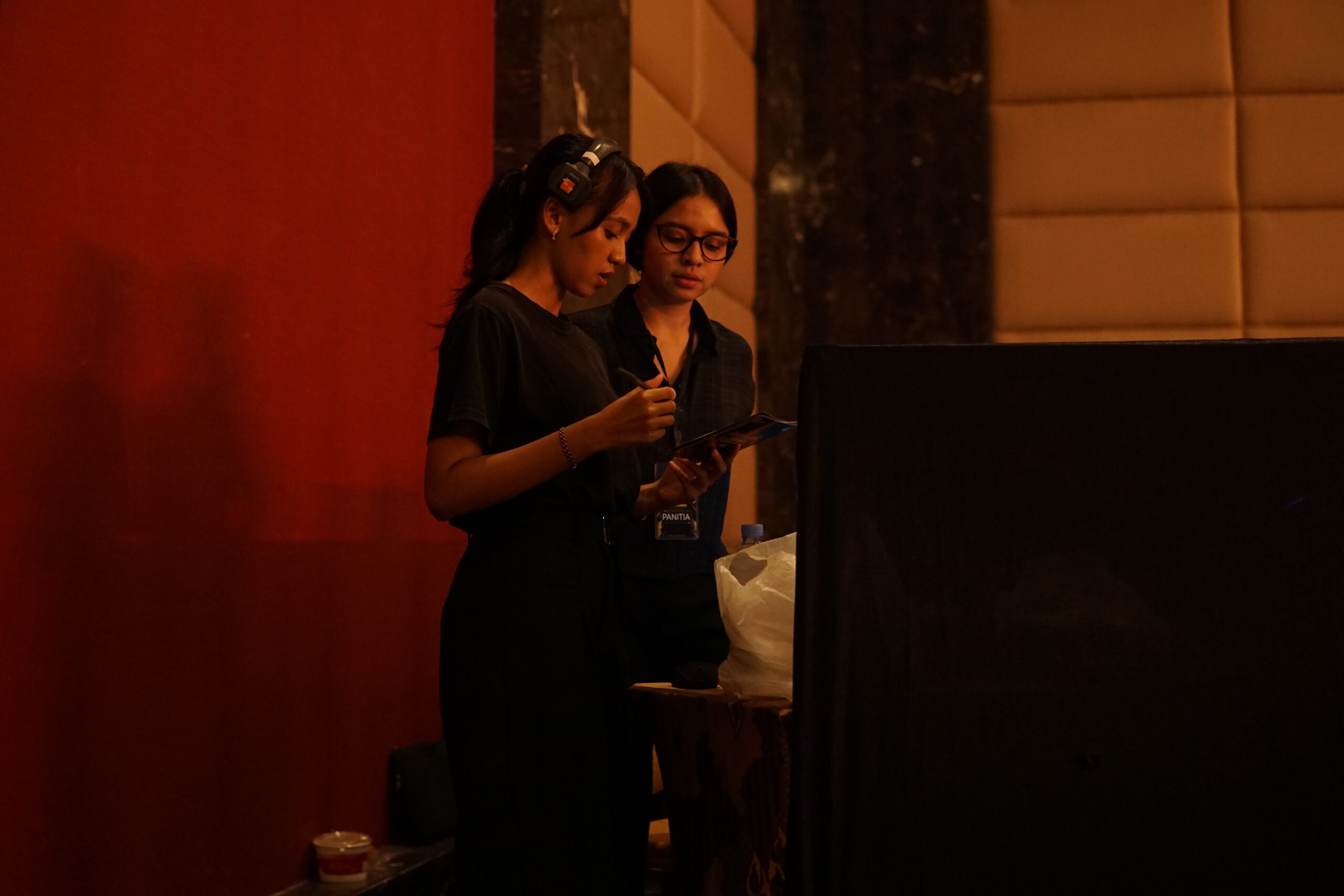 Two event organizers in black attire preparing for an event, reviewing a tablet and holding a microphone, with a red backdrop and modern venue decor in the background