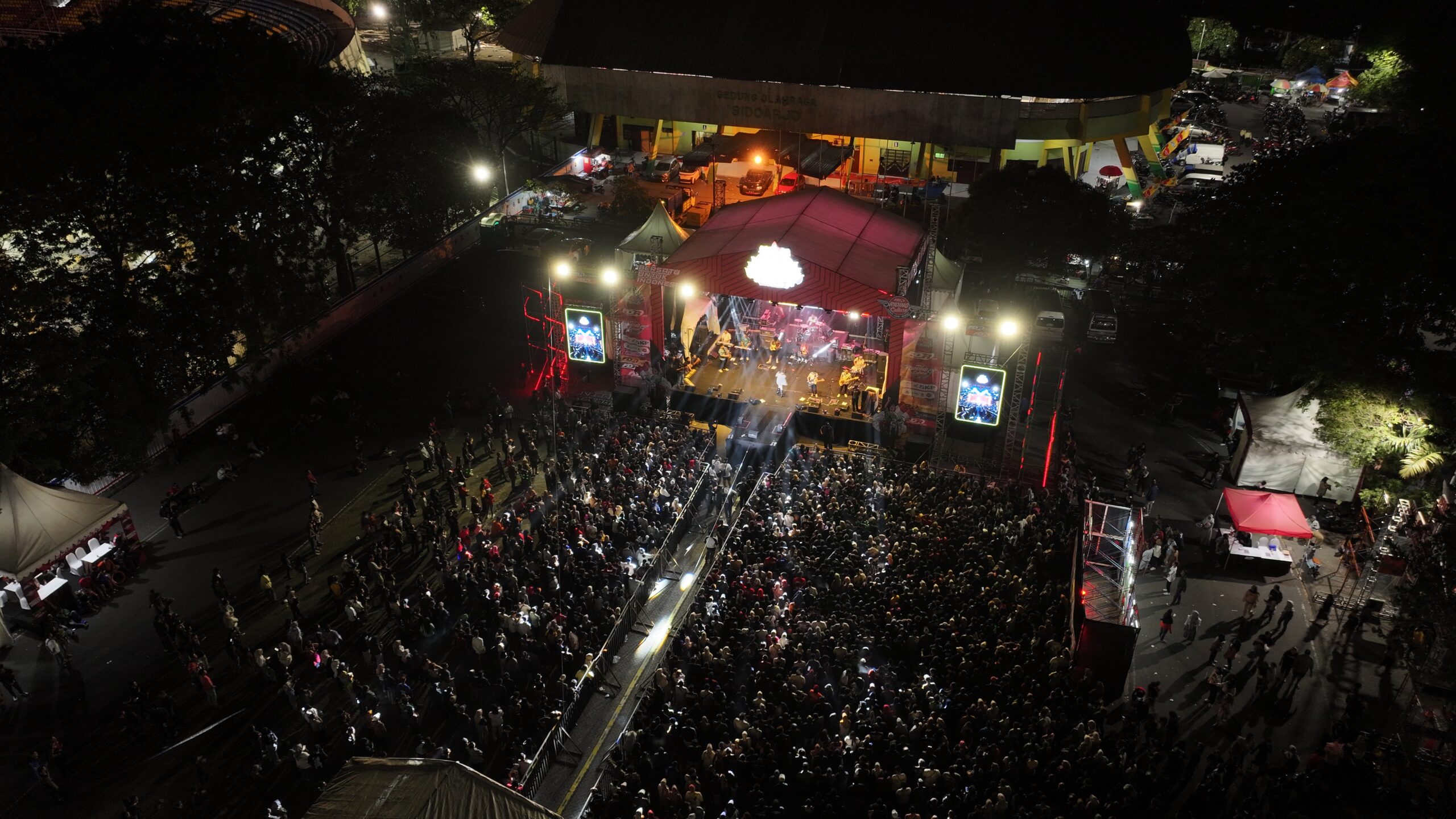 Aerial view of a vibrant outdoor concert at night with a large crowd gathered in front of a brightly lit stage, surrounded by tents, food stalls, and colorful lights, creating a festive atmosphere for an ARIA event.