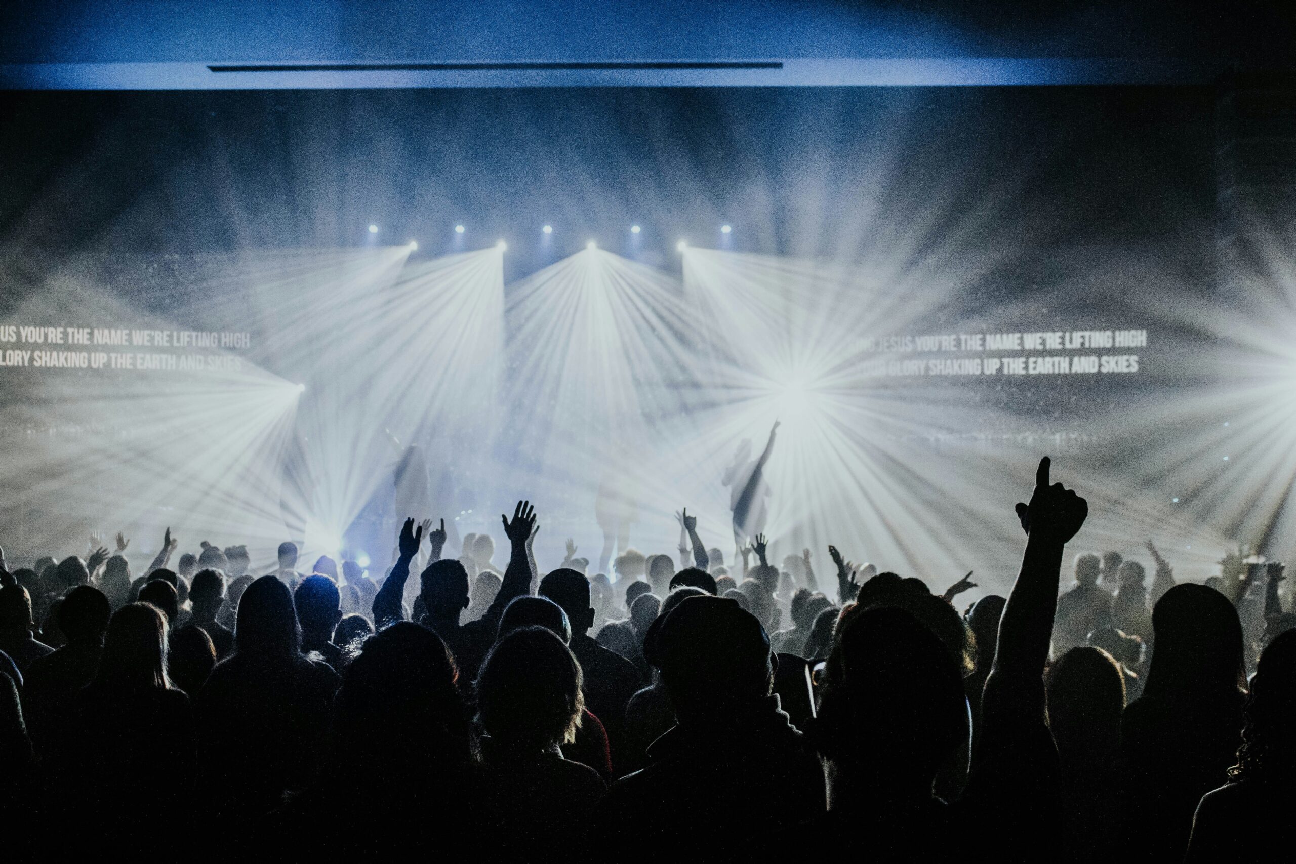 A large crowd at an Aria Event Organizer concert raises their hands in excitement under dramatic stage lighting, with beams of light streaming down and lyrics displayed on a screen in the background.